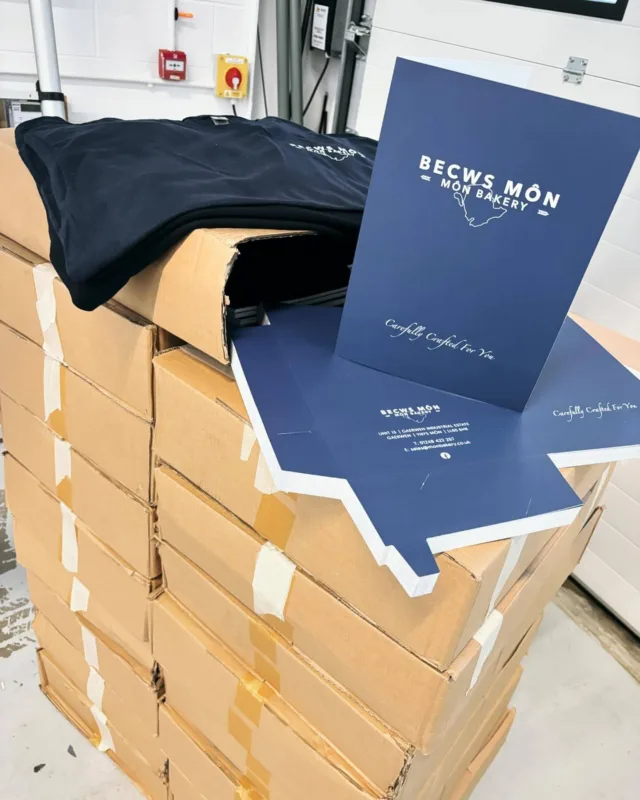 Stacked cardboard boxes with branded BECWS MÔN bakery presentation folders and printed navy t-shirts