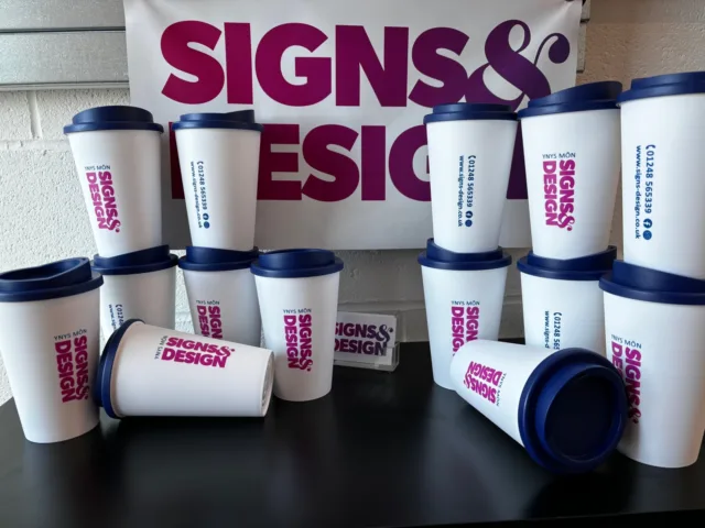 White reusable cups with blue lids featuring purple printed Signs & Design branding, displayed in front of a matching promotional banner